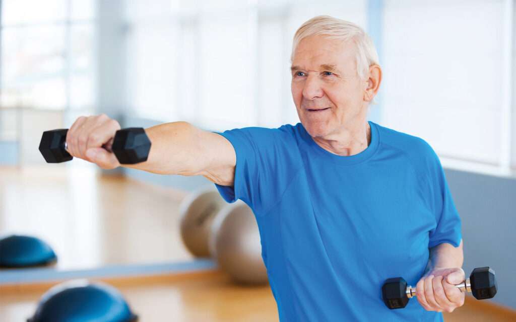 Eine Person in einem Fitnessstudio führt eine Übung mit zwei Kurzhanteln aus. Die Person trägt ein blaues Shirt; der linke Arm ist ausgestreckt, während der rechte angewinkelt bleibt. Im Hintergrund sind Gymnastikbälle und andere Trainingsgeräte sichtbar. Das Bild vermittelt ein aktives, gesundheitsbewusstes Lebensgefühl.