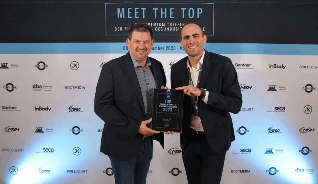 Zwei formell gekleidete Personen halten eine Auszeichnung vor einer Wand mit Logos und dem Schriftzug ‚MEET THE TOP – Der Premium-Treff der Fitness- und Gesundheitsbranche, 09.–12. November 2023‘.